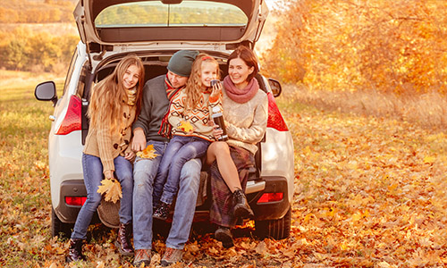 Family of four sitting in the open trunk of a car surrounded by autumn leaves.