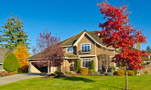 A suburban two-story house with a stone facade, green lawn, driveway, and colorful autumn trees under a clear blue sky.